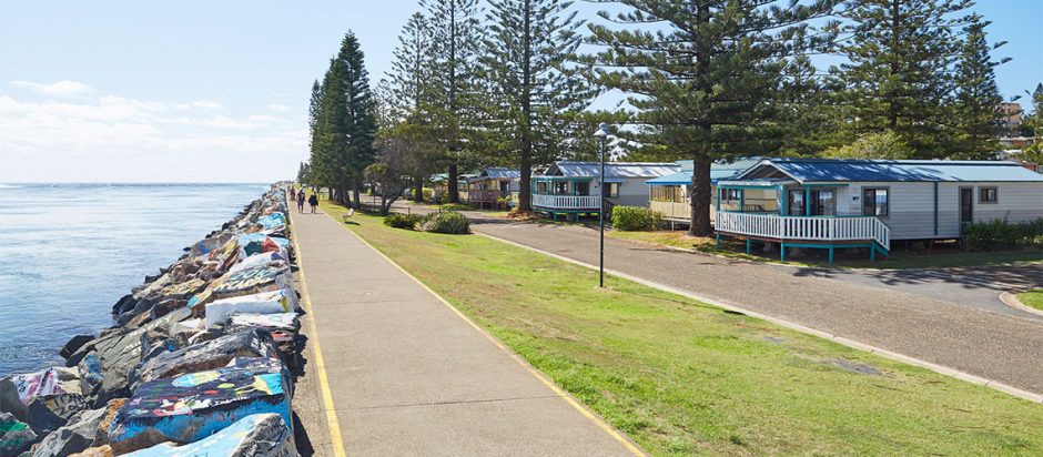 A photo of the NRMA Port Macquarie Holiday Park. It shows a lush green landscape with several holiday cabins visible, suggesting a relaxing and scenic vacation destination.